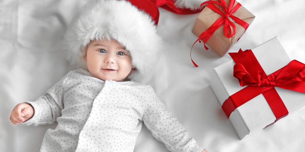 Baby in gepunktetem Schlafanzug liegt mit Weihnachtsmtze auf einer weien Decke neben Geschenken mit roter Schleife