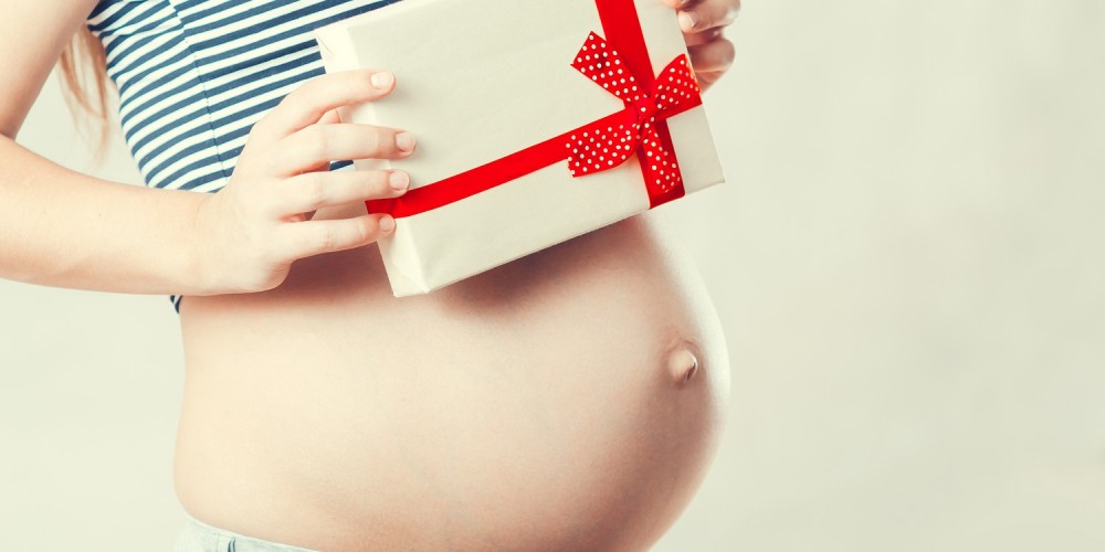 Schwangere Frau mit dickem Babybauch hält ein Geschenk mit rotem Geschenkband in den Händen