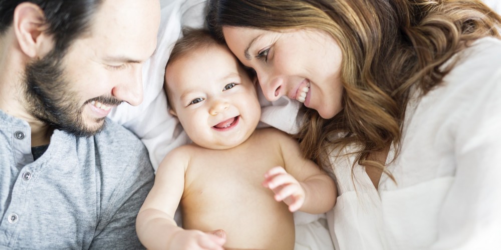 Glückliche Eltern liegen mit ihrem nackten Baby in der Mitte im Bett und lächeln