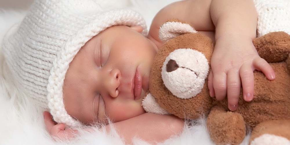 Schlafendes Baby mit Mütze, Neugeborenes mit Teddy im Arm auf Fell