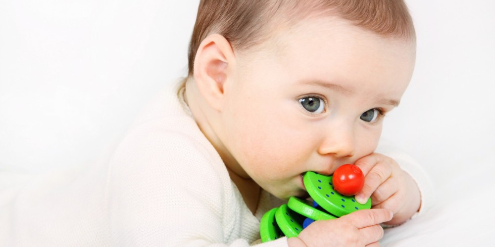 Dunkelhaariges Baby nuckelt an einem grnen Holzspielzeug fr Babys