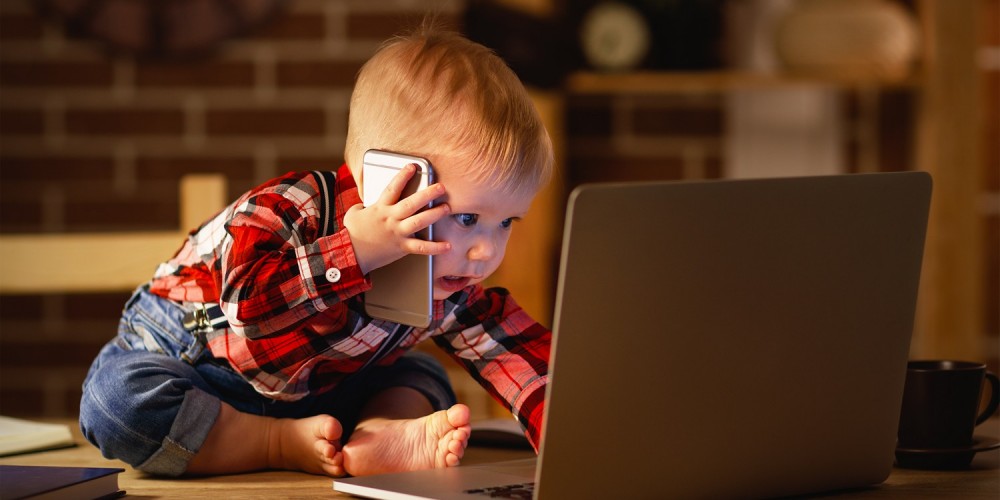 Kleiner Junge in rotem Karohemd und Jeans sitzt vor einem Laptop auf dem Tisch und hlt ein Smartphone ans Ohr