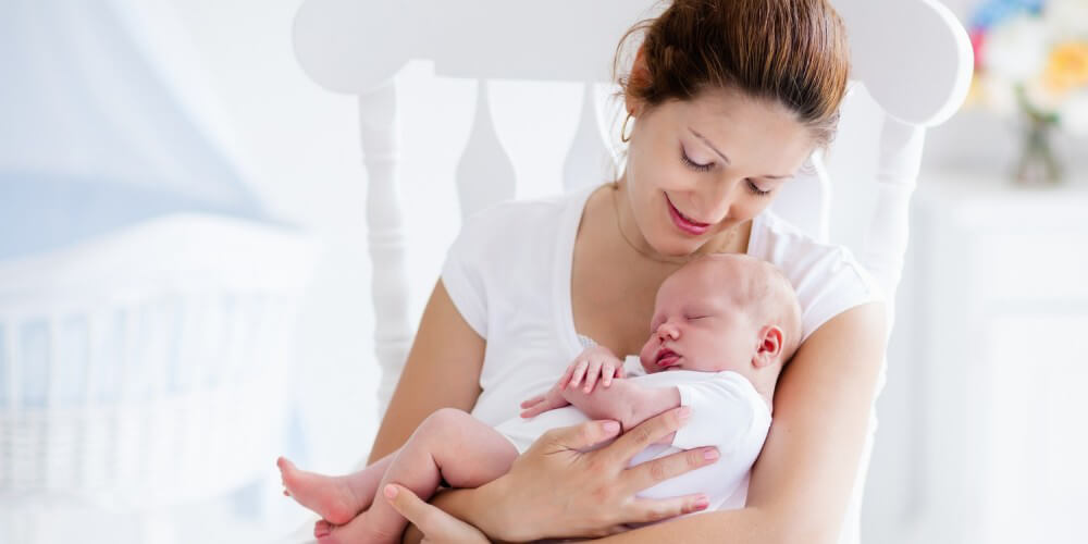 Mutter sitzt mit ihrem Baby im Arm in einem weien Schaukelstuhl im Babyzimmer