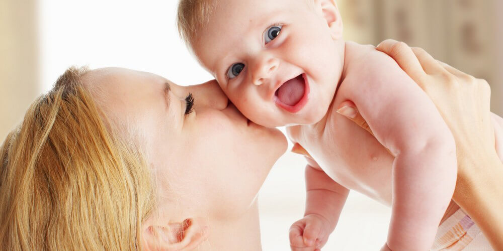 Blonde Mutter hält ihr fröhlich guckendes Baby hoch und küsst es auf die Wange
