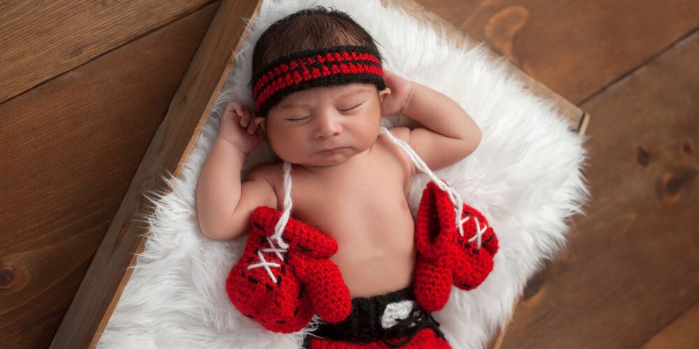 Baby liegt in gestrickten Boxer-Bekleidung und mit roten Woll-Boxhandschuhen auf einem Lammfell