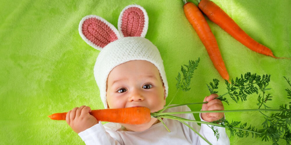 Baby liegt als Hase verkleidet neben zwei Mhren auf einer grnen Decke und beit in eine Mhre