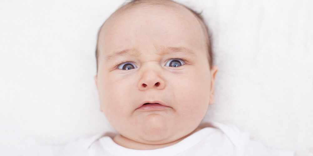 Dunkelhaariges Baby in weißem Body liegt auf weißer Decke und guckt verärgert in die Kamera