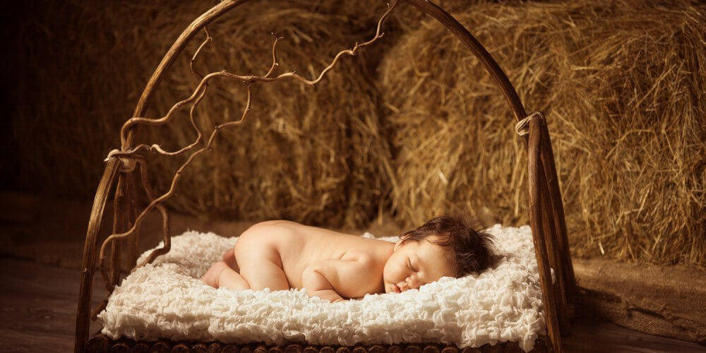 Baby liegt nackt schlafend in einer Krippe aus Holz, im Hintergrund liegen Strohballen