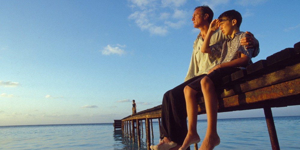 Vater und Sohn sitzen auf Bootssteg am Meer, schauen in den Sonnenuntergang, blauer Himmel