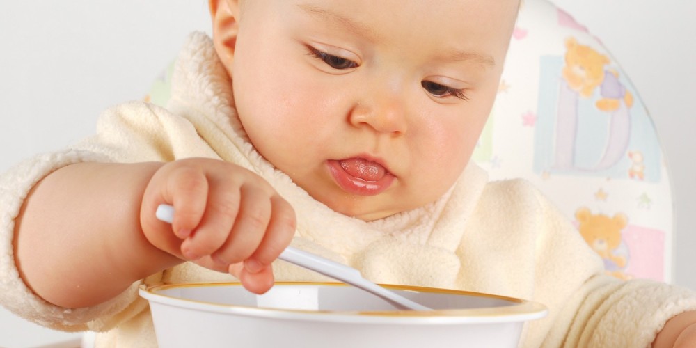 Baby im gelben Strampler isst mit Plastiklffel aus Plastikschale, die Zunge halb herausgesteckt