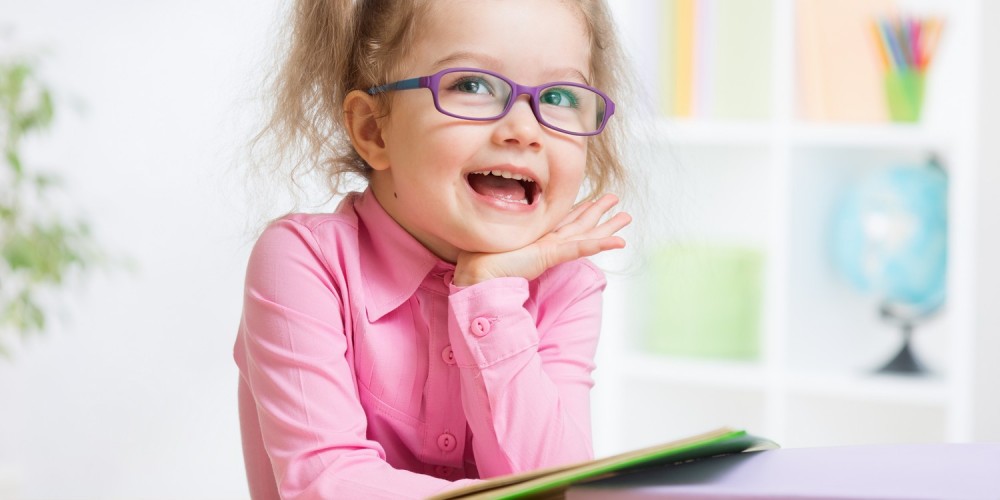 Kleines Mdchen mit Zpfen und lila Brille sitzt mit Bchern an einem Tisch und trumt vor sich hin