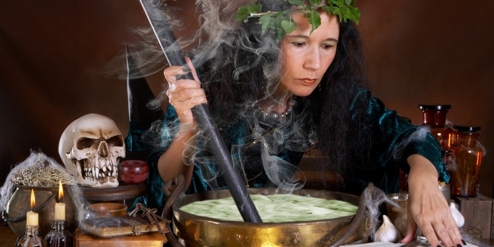 Frau als Hexe verkleidet rührt in grüner Flüssigkeit rum, umgeben von Totenkopf, Kerzen etc.
