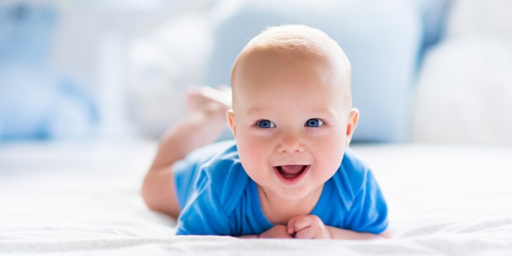 Lächelnder Babyjunge krabbelt in blauem Strampler auf einem Bett