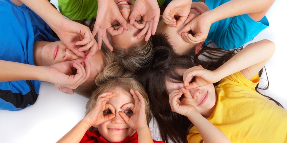 Fnf Kinder liegen im Kreis auf dem Boden, halten Hnde um Augen und schauen hoch in Kamera